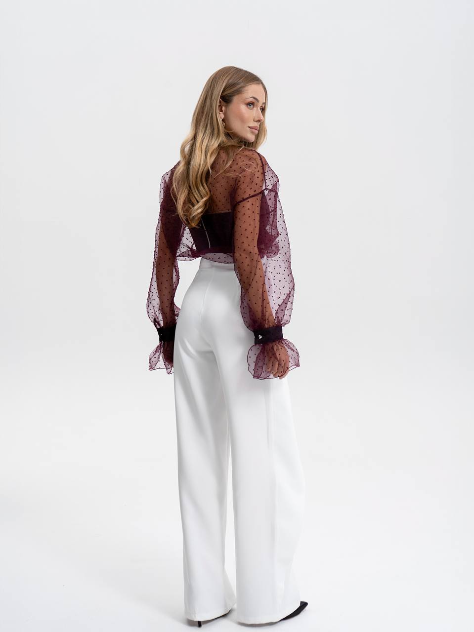 Woman wearing a sheer burgundy top and white pants on a white background