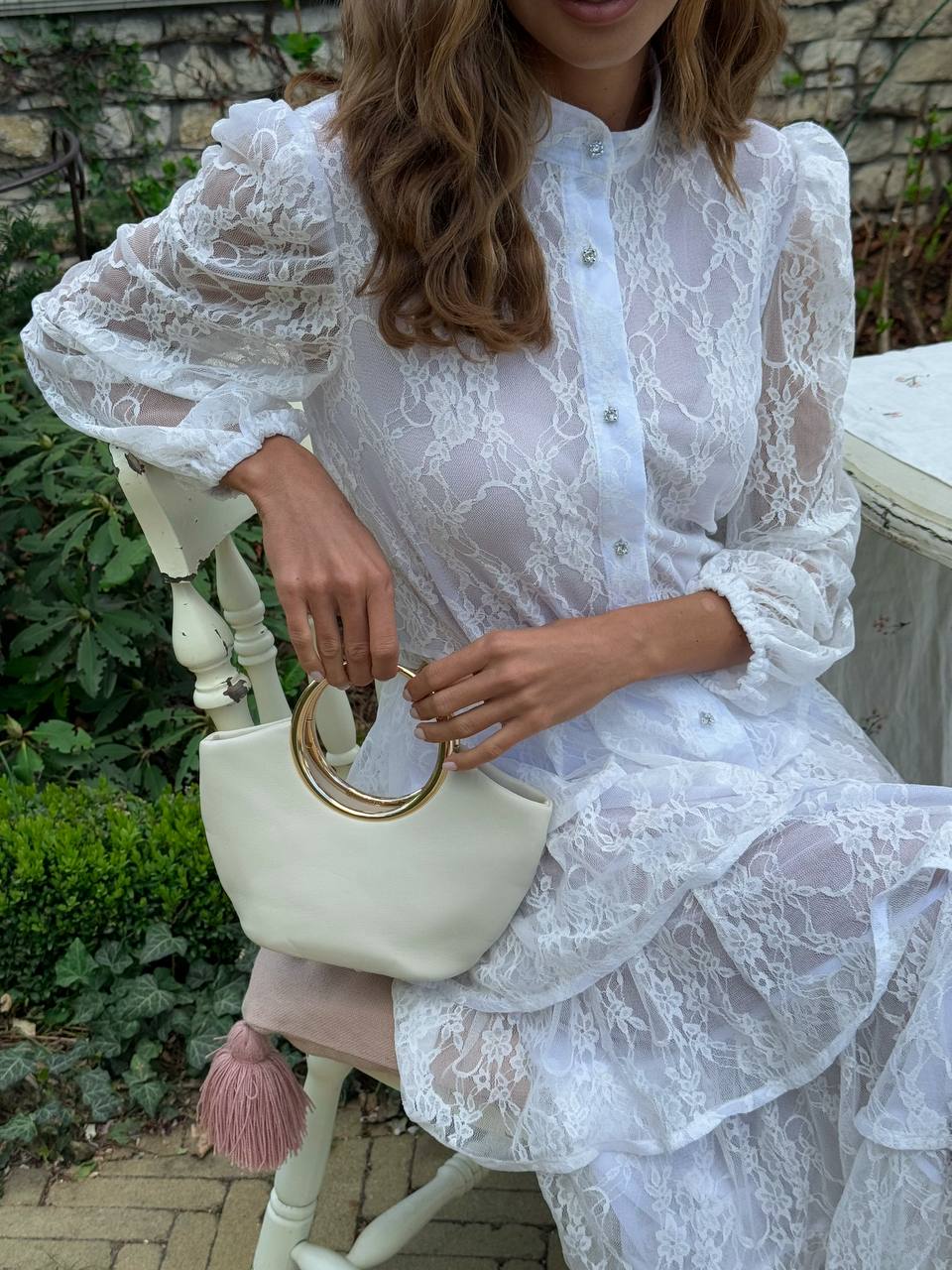 Woman in a white lace dress holding a cream handbag with a pink tassel outdoors.