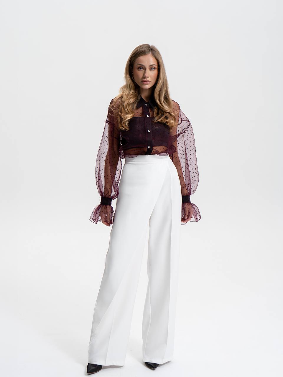 Woman wearing a dark blouse with sheer sleeves and white pants on a white background