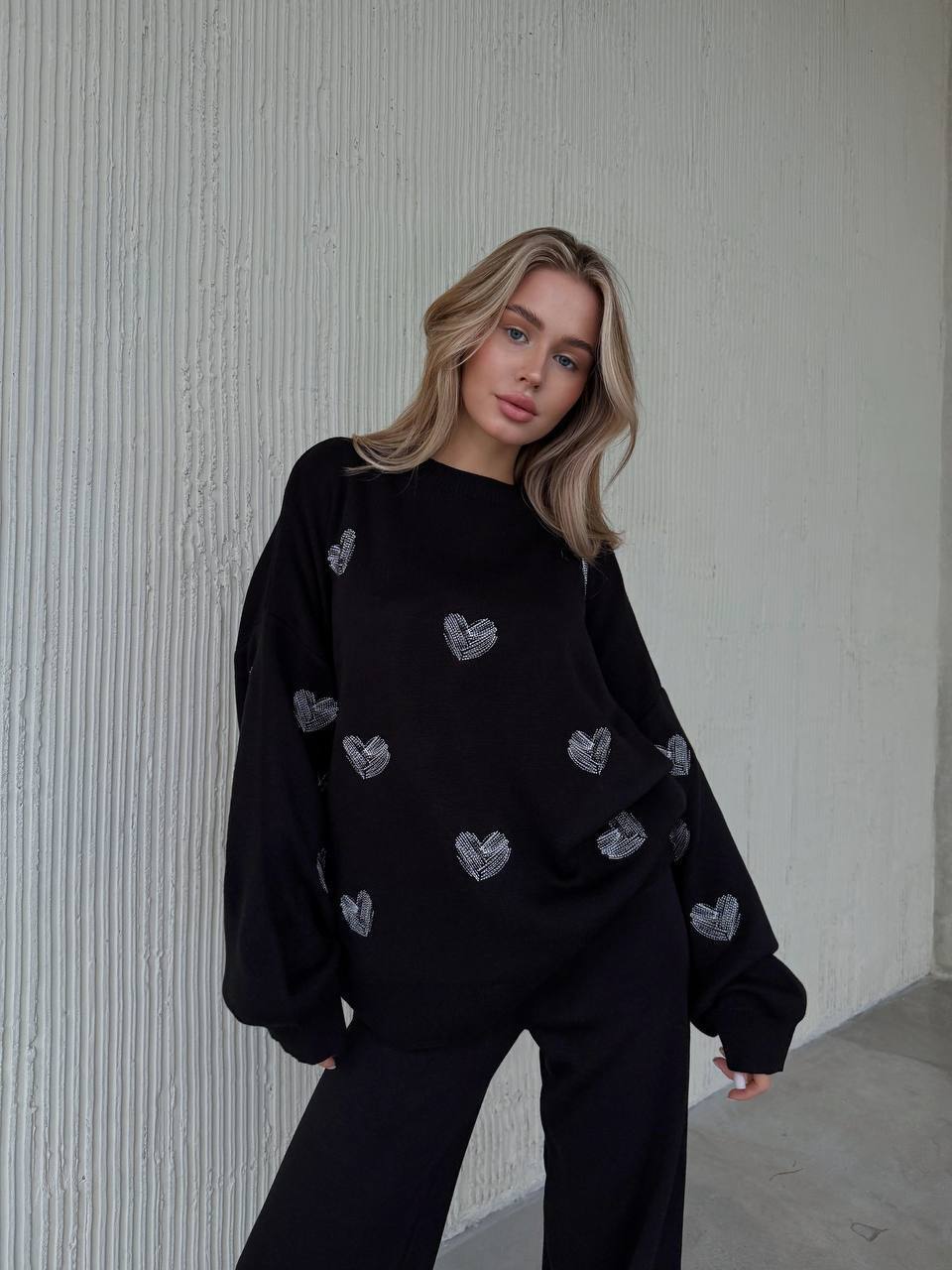 Woman wearing a black sweater with heart patterns against a textured white wall.