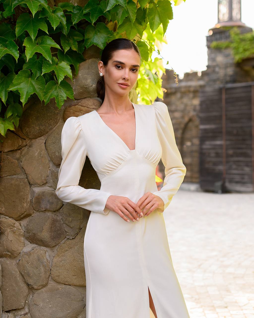 Woman in a white dress standing against a stone wall with greenery