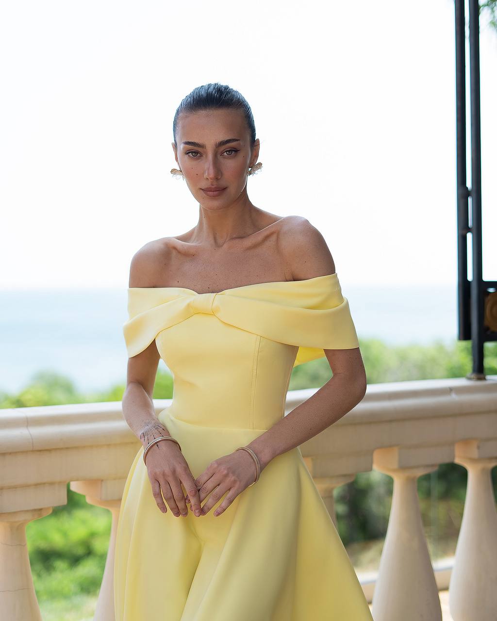 Woman in a yellow off-shoulder dress standing outdoors with greenery in the background