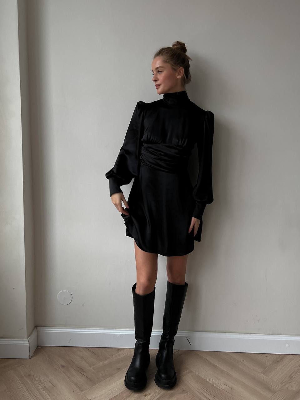 Person wearing a black outfit with a high collar and knee-high boots against a plain wall.