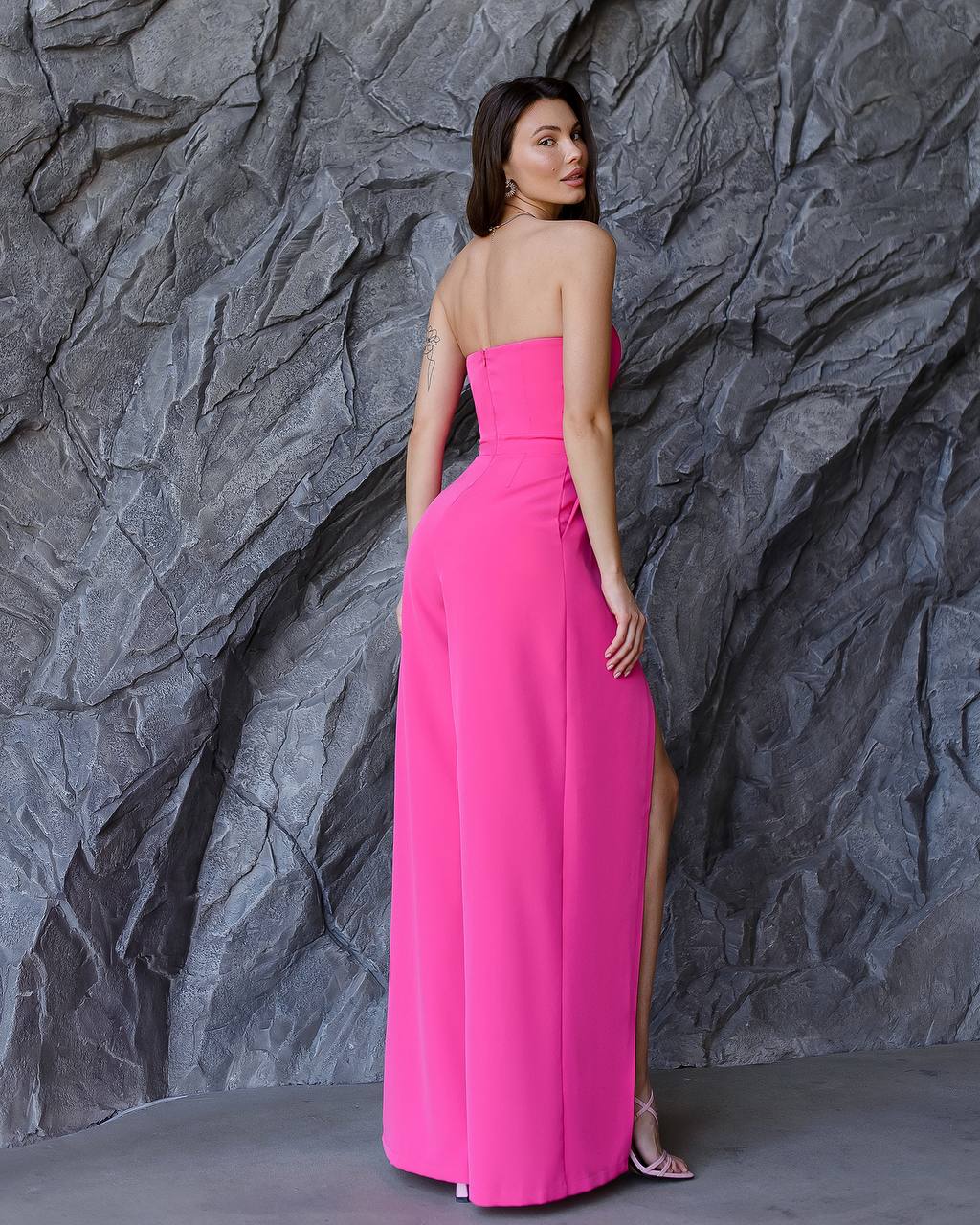 Woman in a pink strapless gown standing against a textured gray wall.
