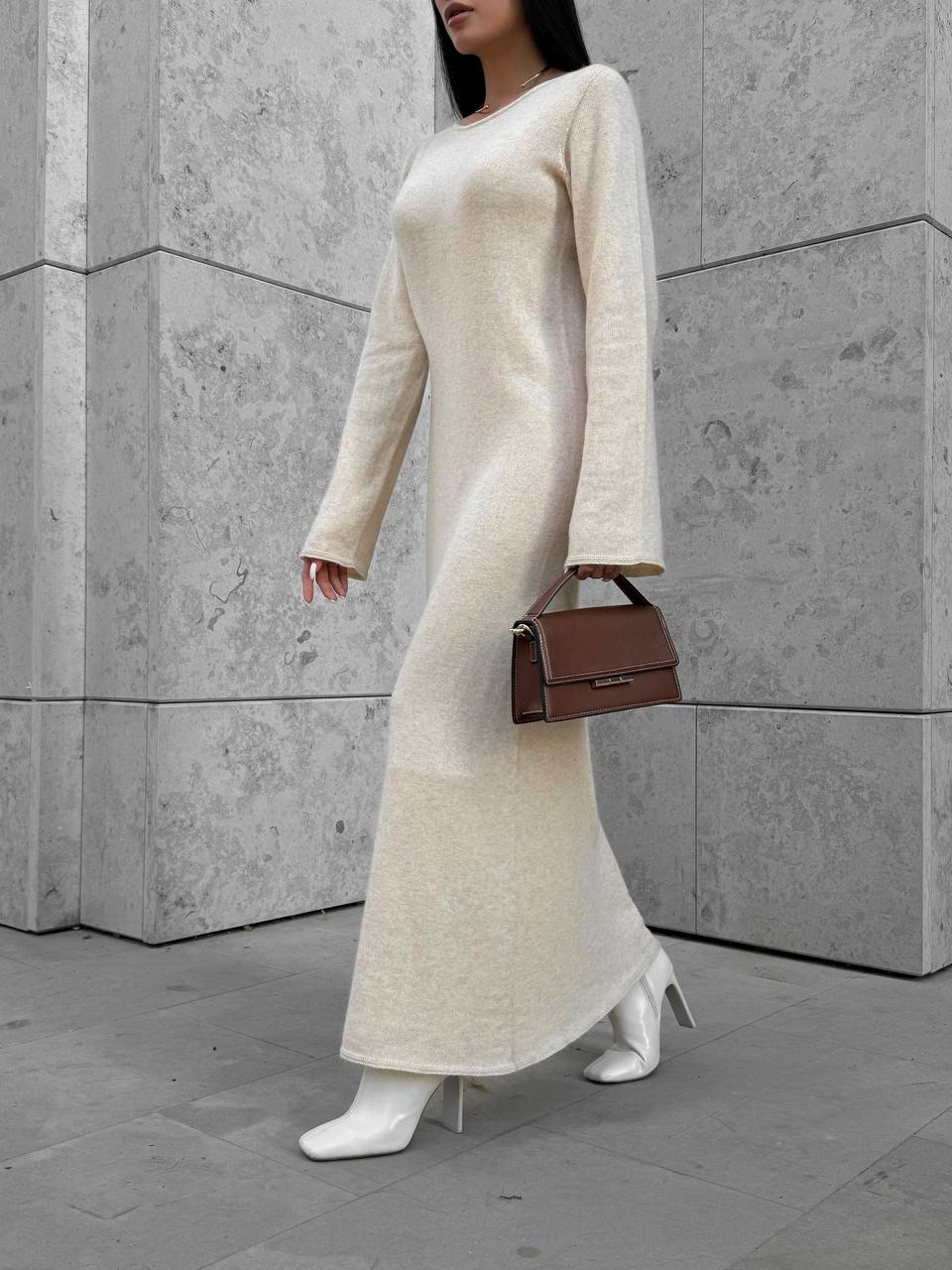Person wearing a long cream knit dress with white boots and a brown handbag against a gray concrete wall.