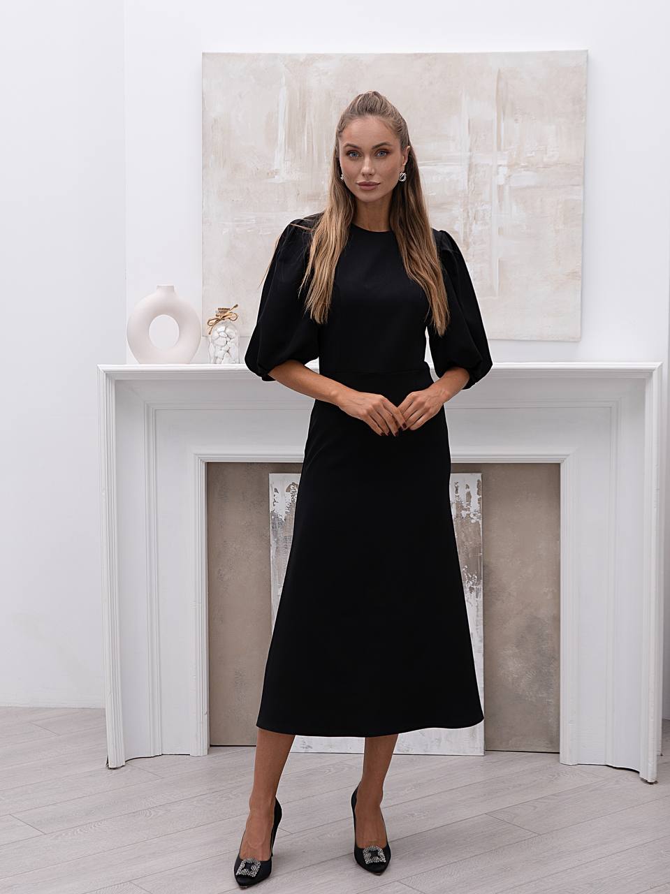 Woman wearing a black dress standing in a room with a white fireplace and abstract painting.