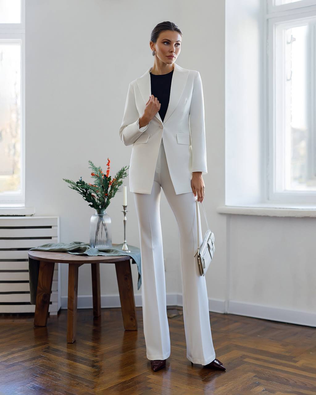 Woman in a white suit standing in a room with wooden floor and large windows.