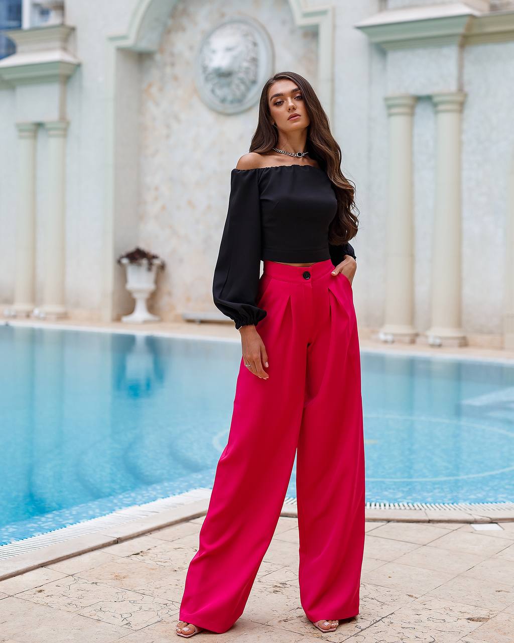 Woman in black top and pink pants standing by a pool with classical architecture in the background
