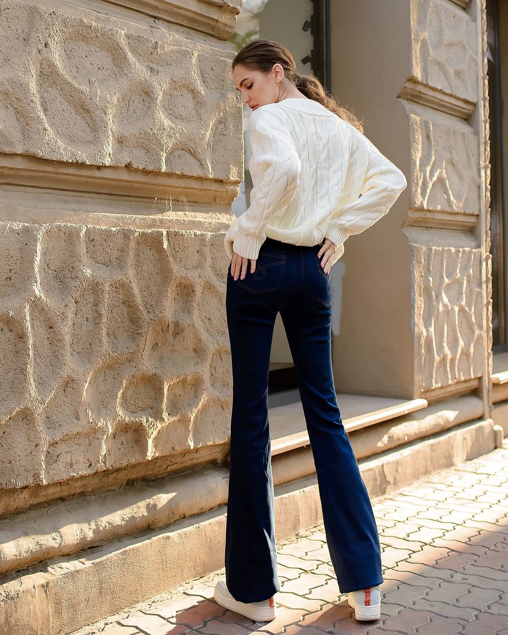 Woman in a white blouse and blue flared pants standing against a stone wall.