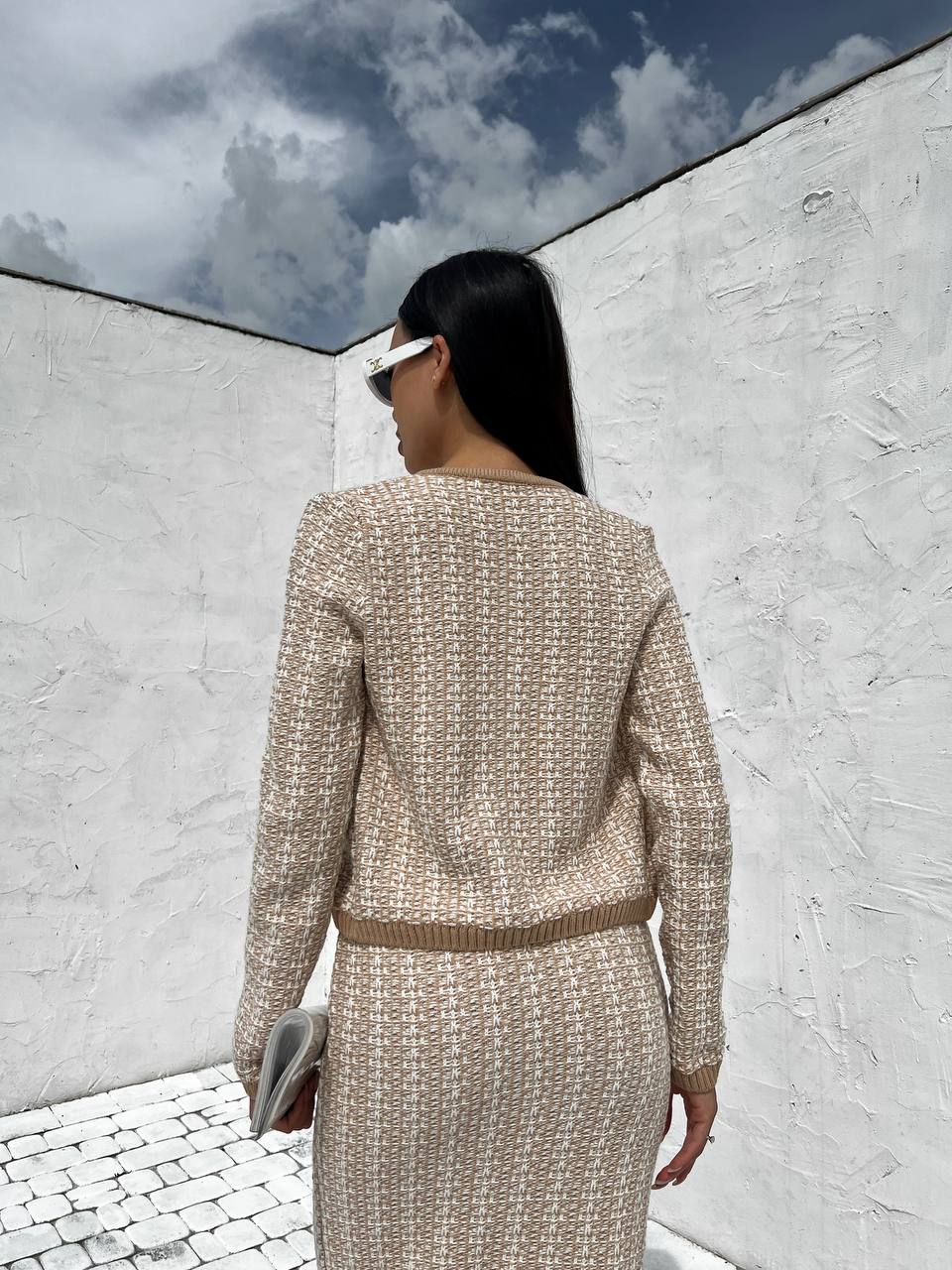 Woman in a beige tweed suit standing against a white wall with blue sky.