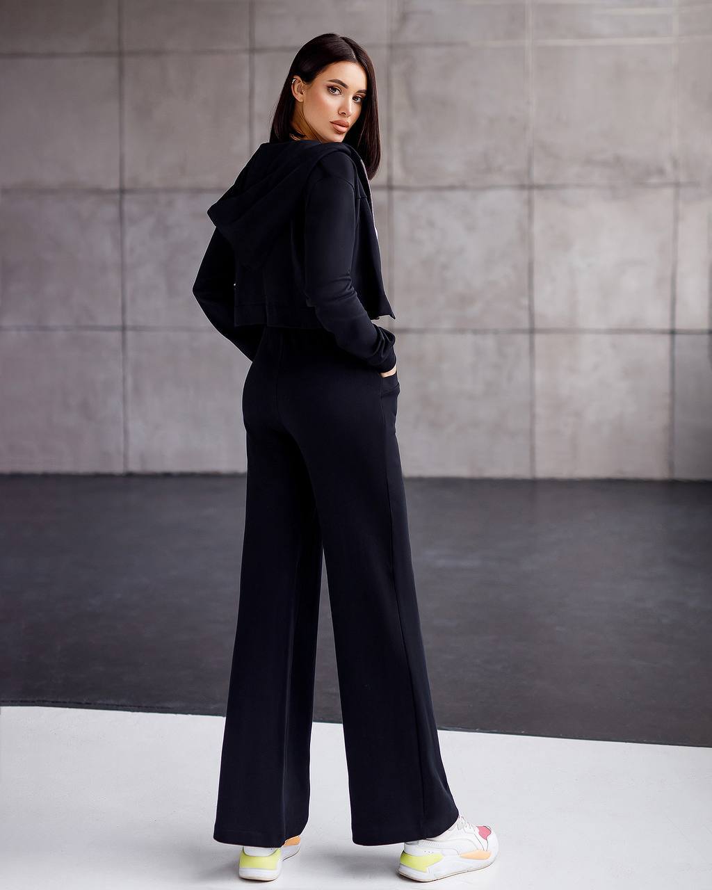 Woman wearing a black outfit with wide-leg pants against a gray wall.
