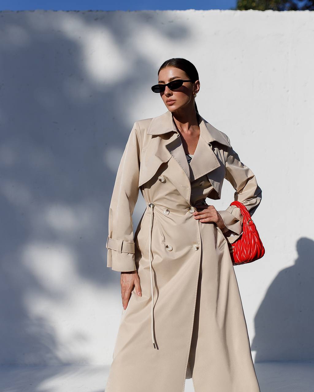 Woman wearing a beige trench coat with a red handbag against a white wall.