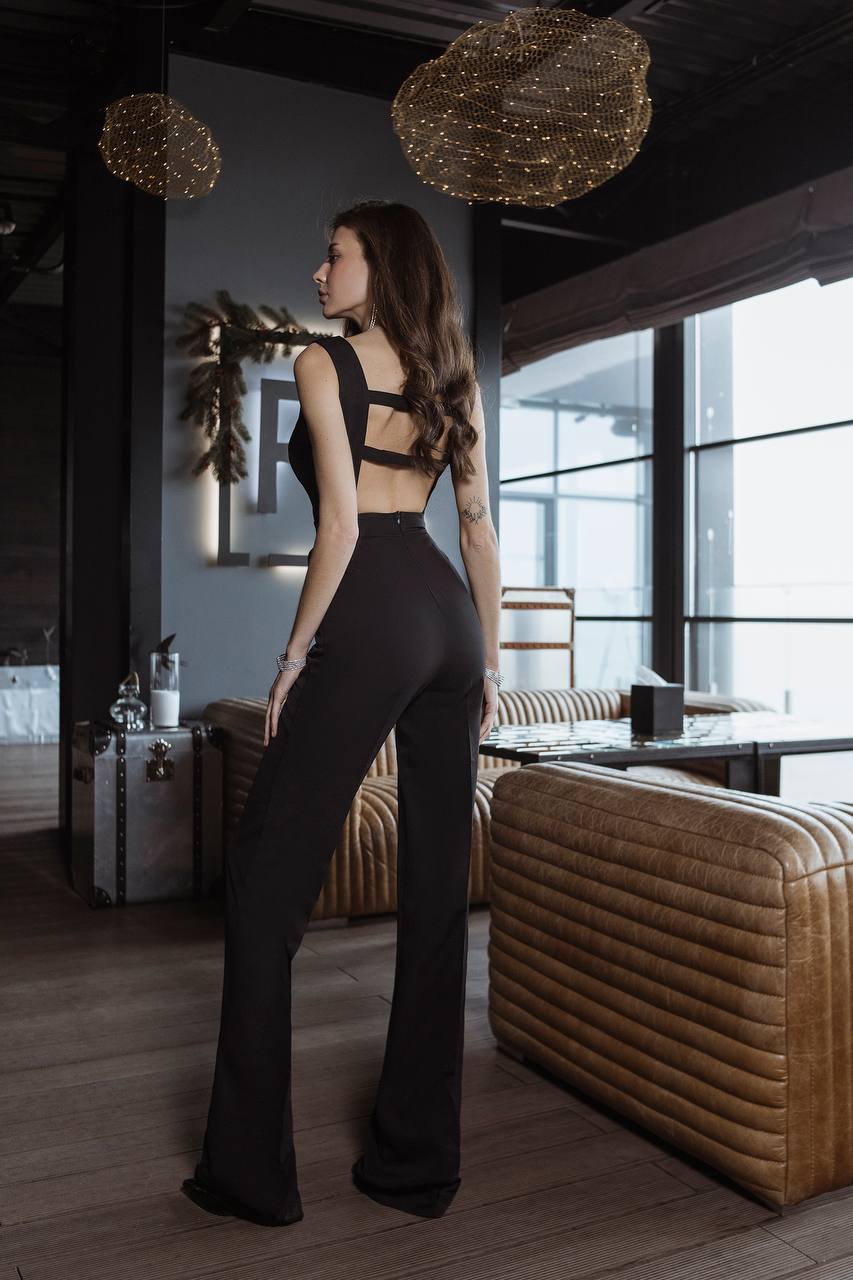 Woman in black outfit standing in a modern interior setting with large windows and decorative lights.