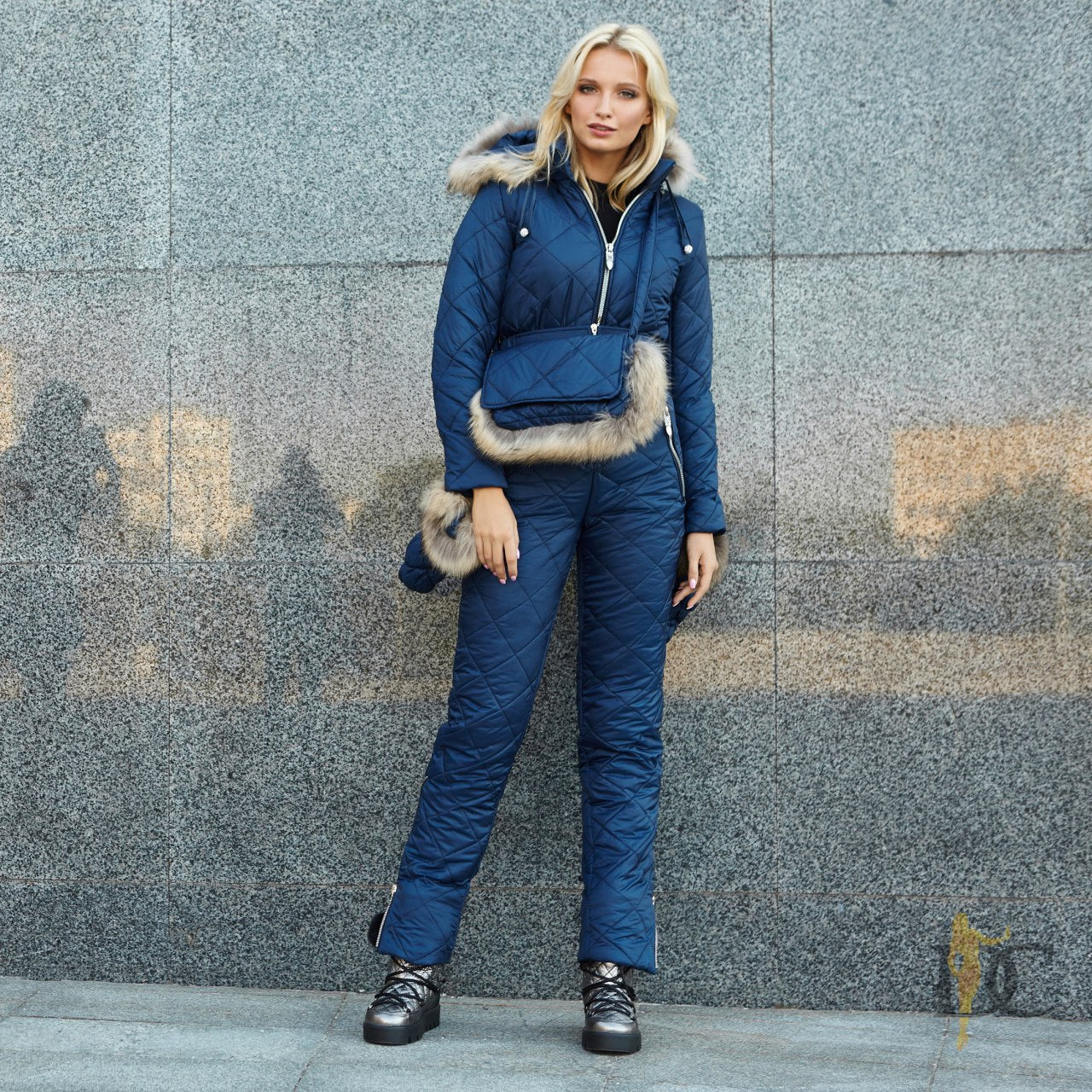 Woman wearing a blue winter outfit with fur-lined hood against a gray wall.