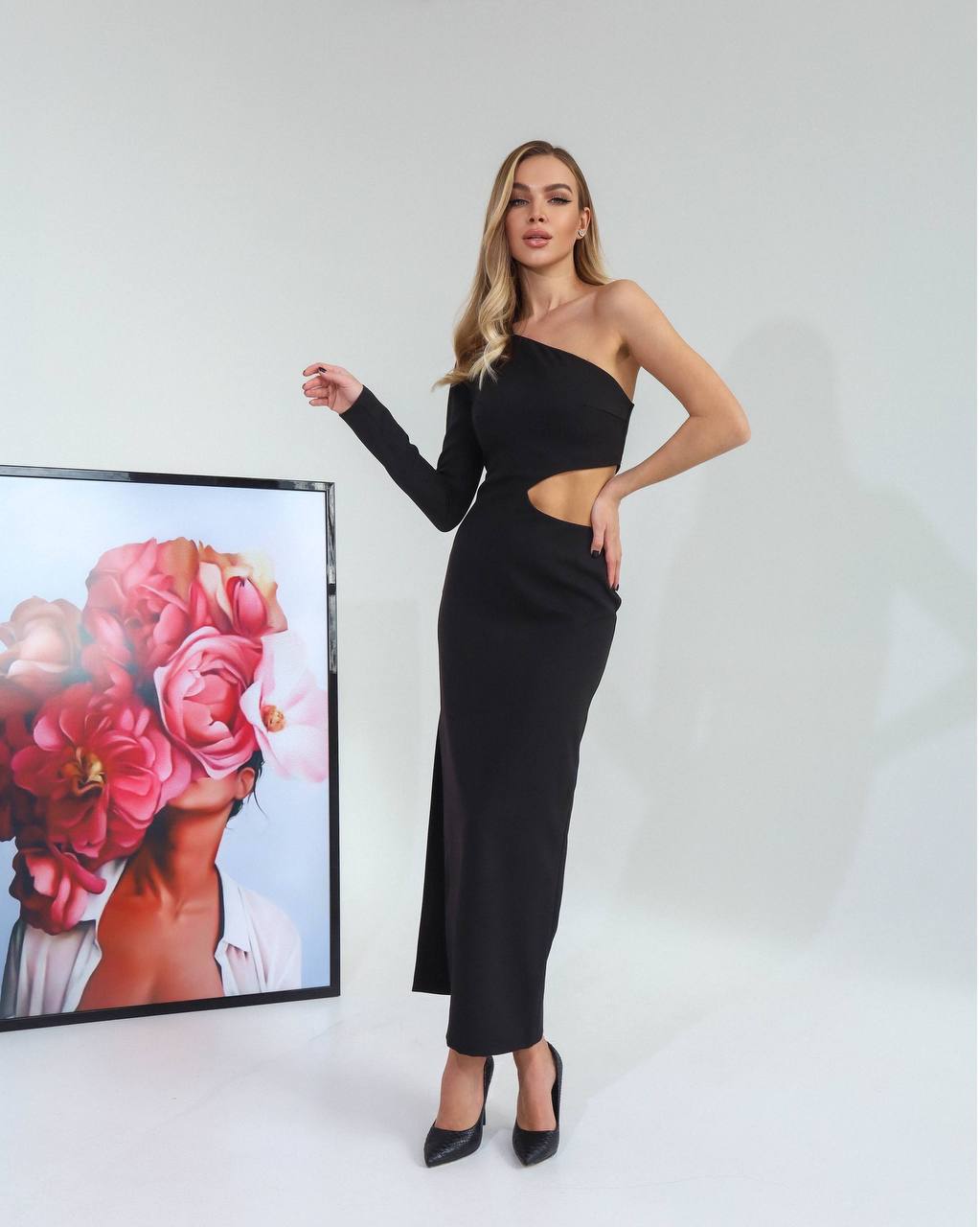 Woman wearing a black one-shoulder dress standing next to a framed picture of flowers.