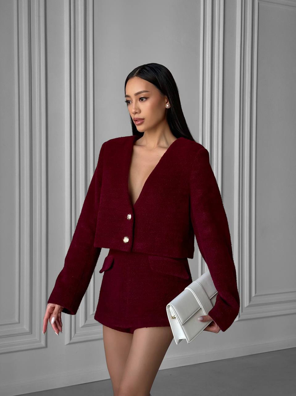 Woman in a burgundy outfit holding a white clutch against a gray wall.