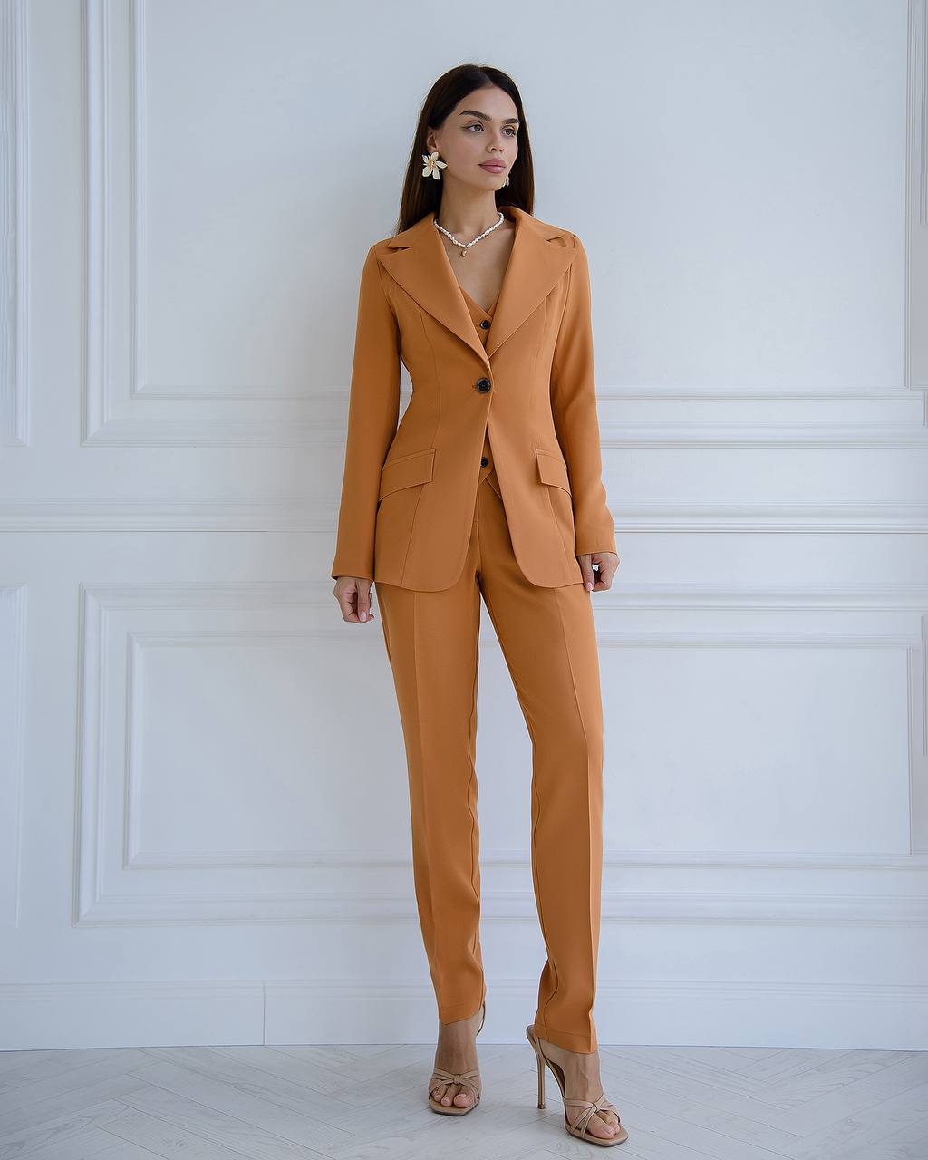Woman wearing a mustard yellow suit against a white paneled wall.