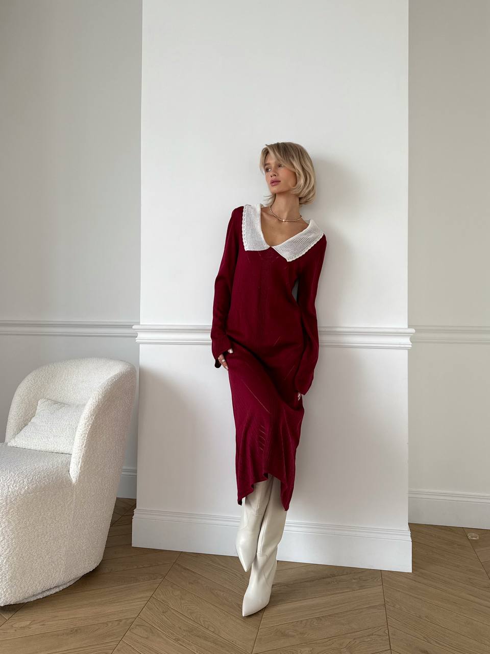 Woman in a burgundy dress with white collar standing against a white wall.