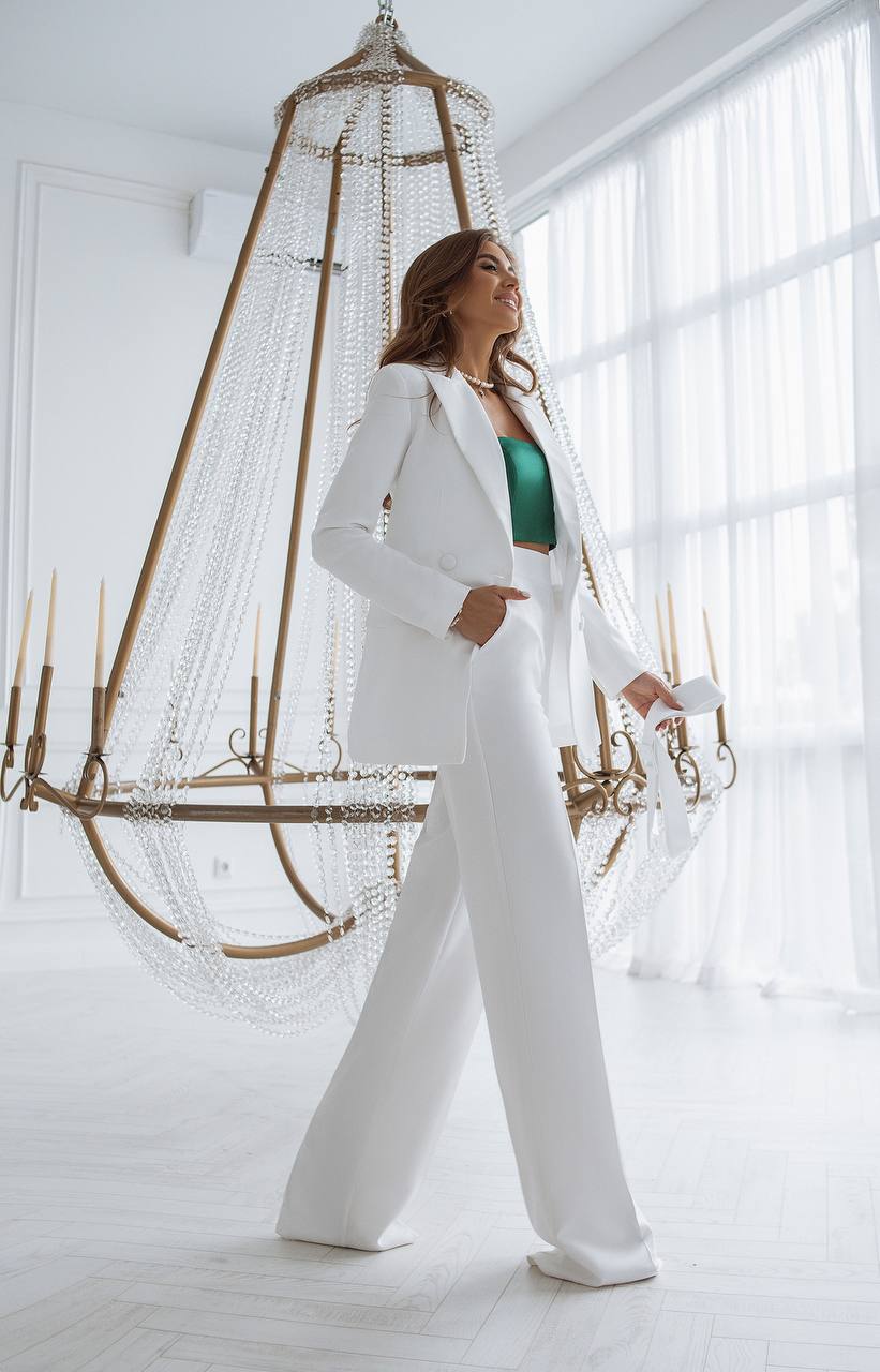 Woman in a white suit standing in a bright room with large windows and a decorative chandelier.