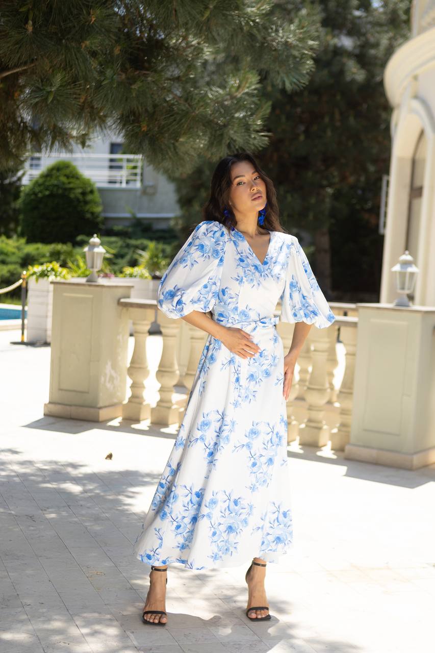 Woman in a blue floral dress standing outdoors with greenery and architectural elements in the background