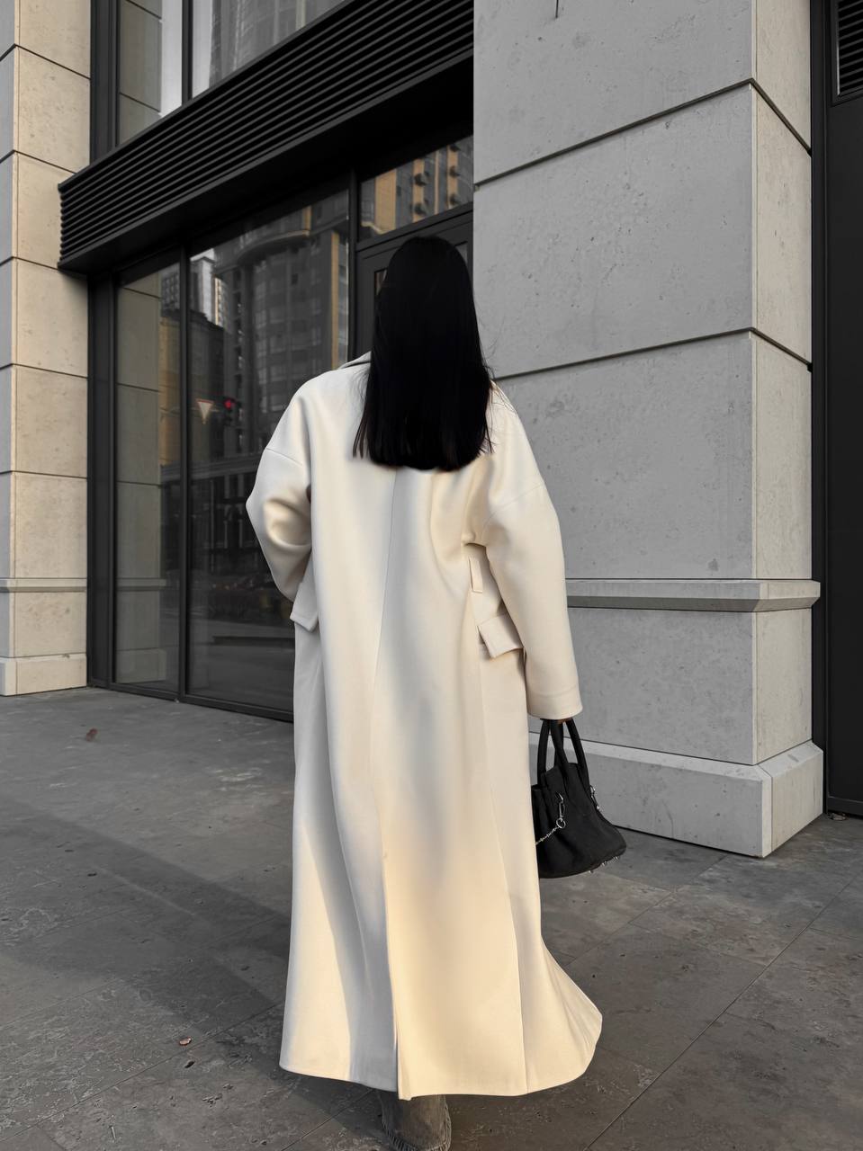 Person wearing a long cream coat walking past a building entrance.
