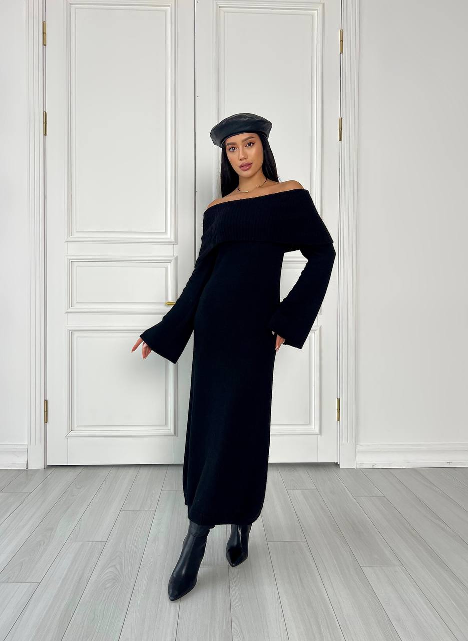 Woman wearing a black off-shoulder dress and beret standing against a white paneled wall.