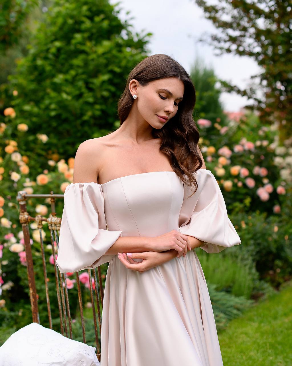 Woman in a white off-shoulder dress standing in a garden with flowers and greenery.