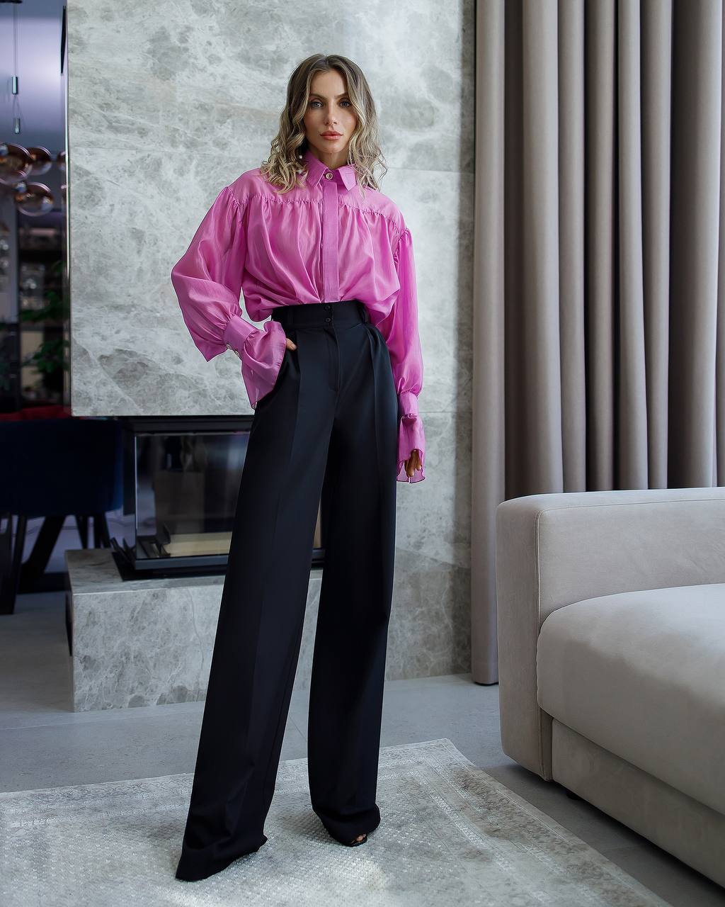 Woman wearing a pink blouse and black pants in a modern living room.