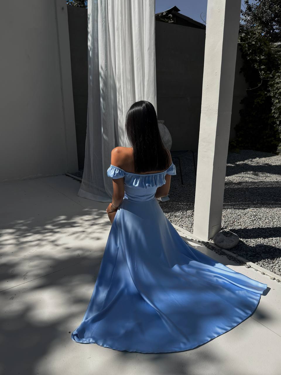 Woman in a blue dress standing in a sunlit outdoor area with white curtains.