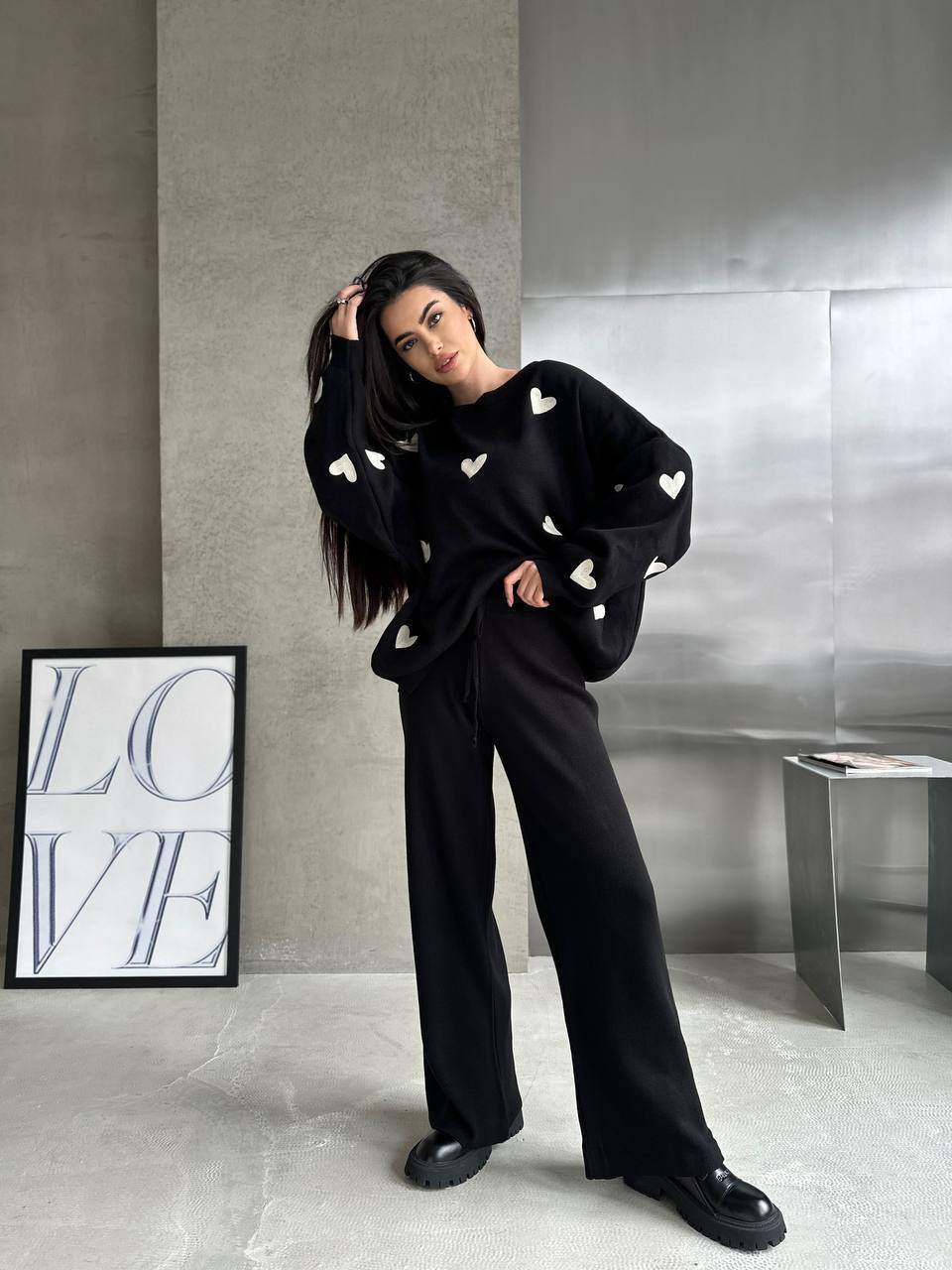 Person wearing a black sweater with white heart patterns and black pants in a modern interior setting.