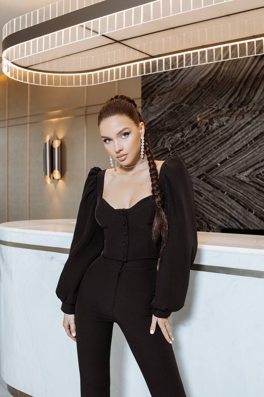 Woman in a black outfit standing in a modern interior setting with a decorative wall and light fixture.
