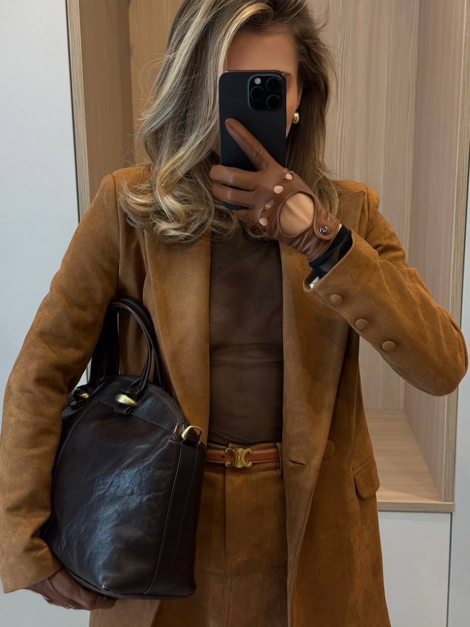 Person wearing a brown coat holding a black bag, taking a mirror selfie.