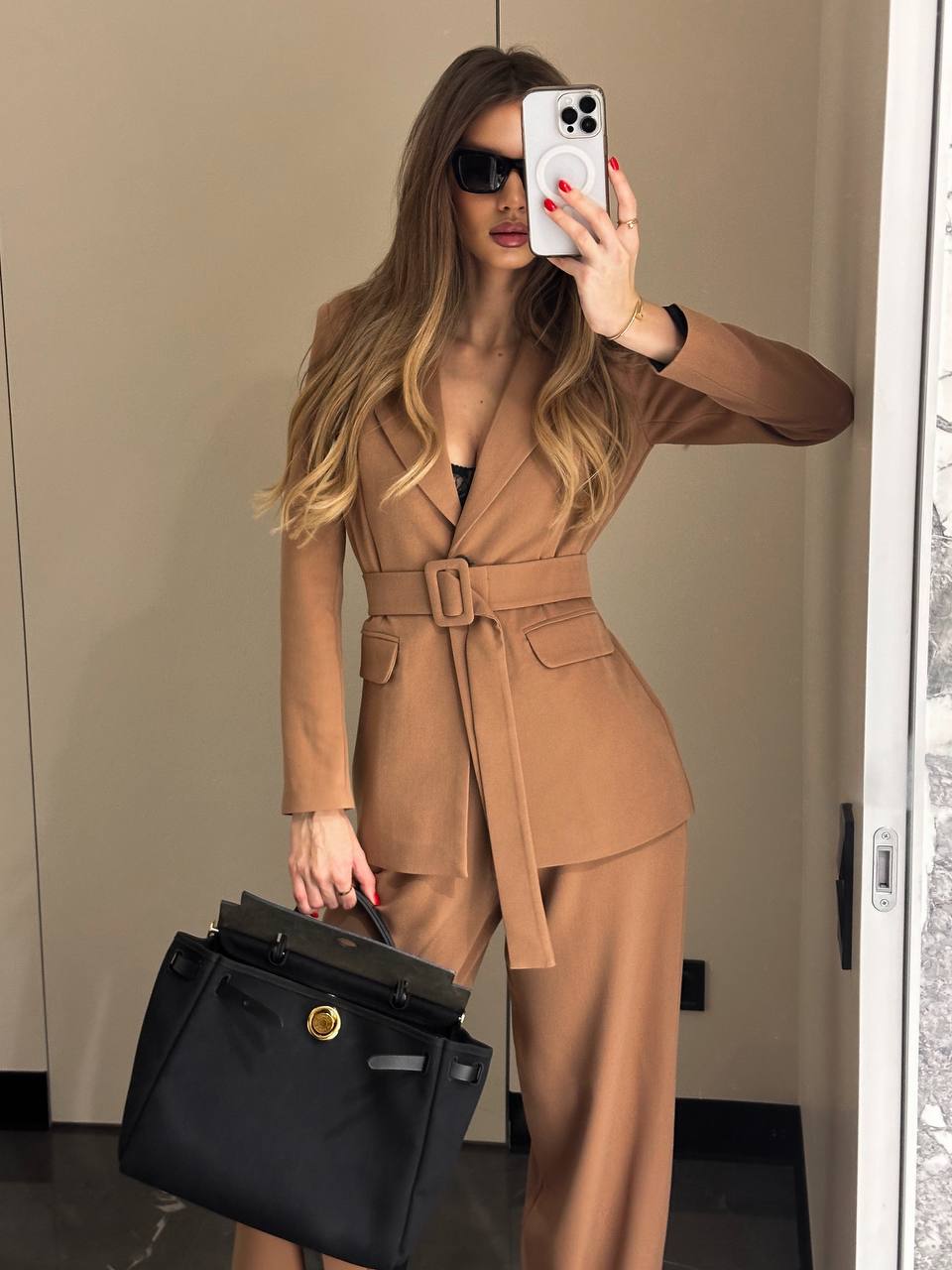 Woman in a brown suit holding a black bag, taking a mirror selfie.