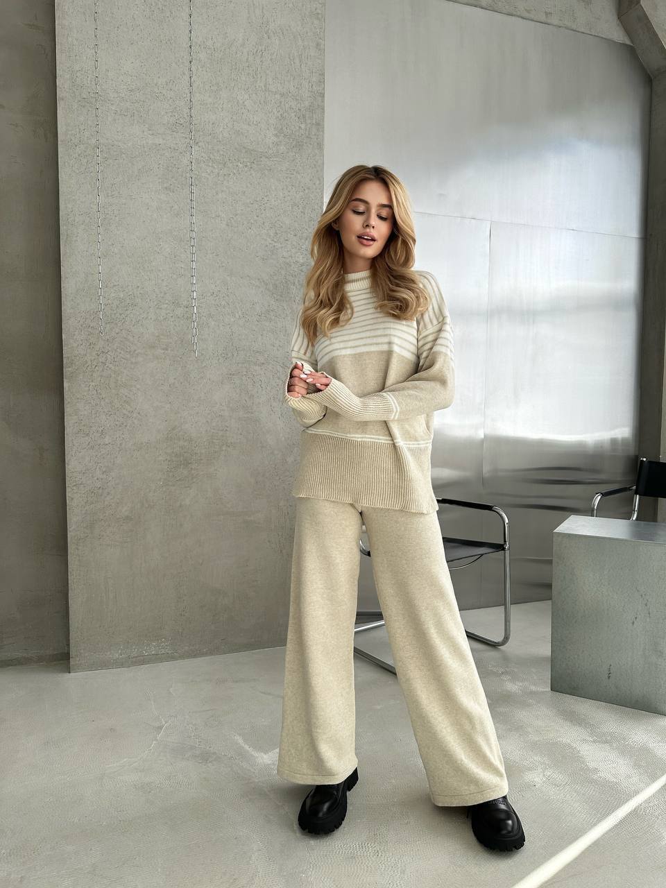 Woman in a beige sweater and pants standing in a modern indoor setting.