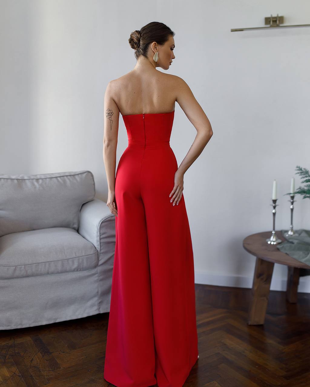 Woman wearing a red strapless jumpsuit in a room with a gray sofa and wooden floor.