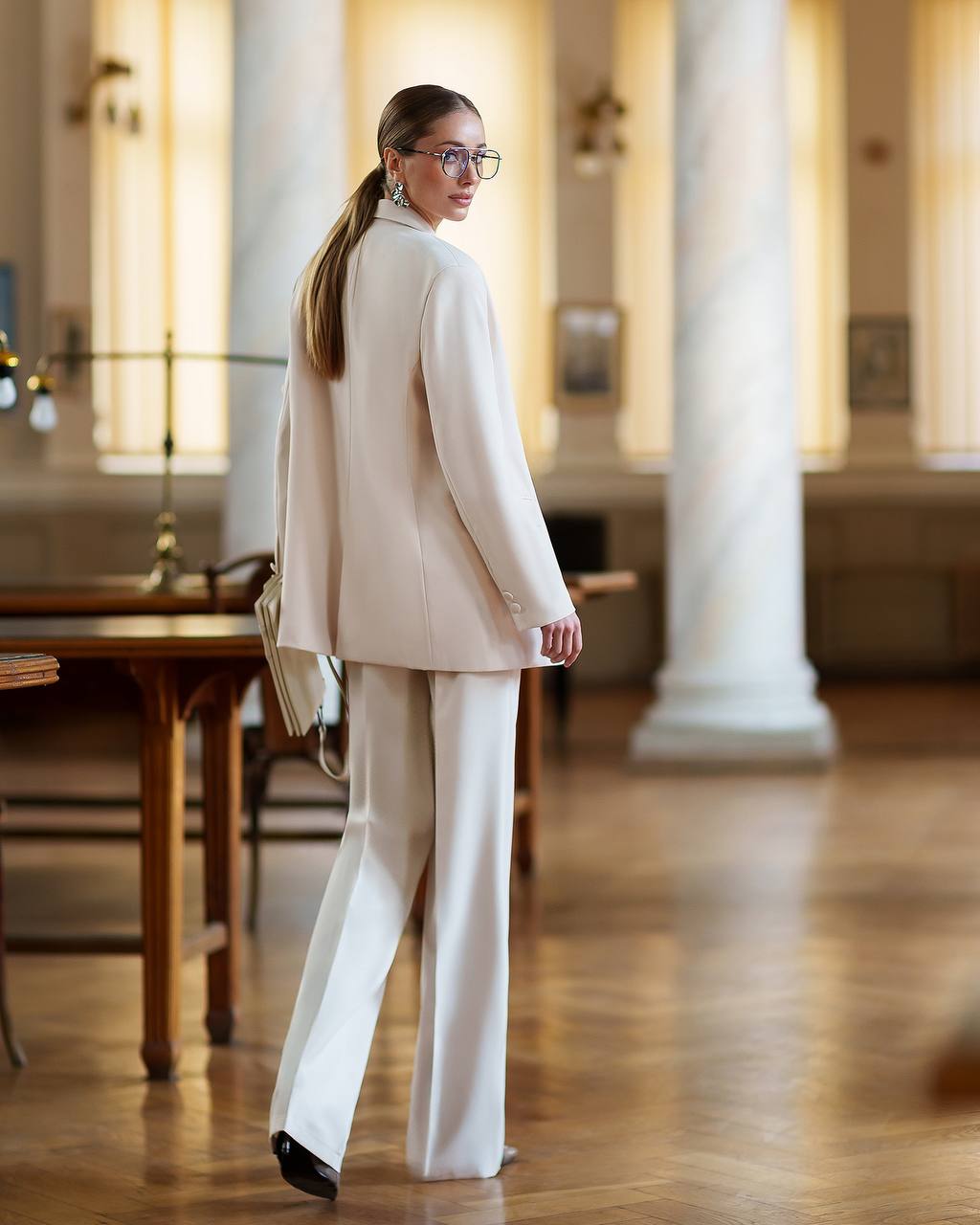 Woman in a beige suit standing in an elegant room with wooden floors and columns.