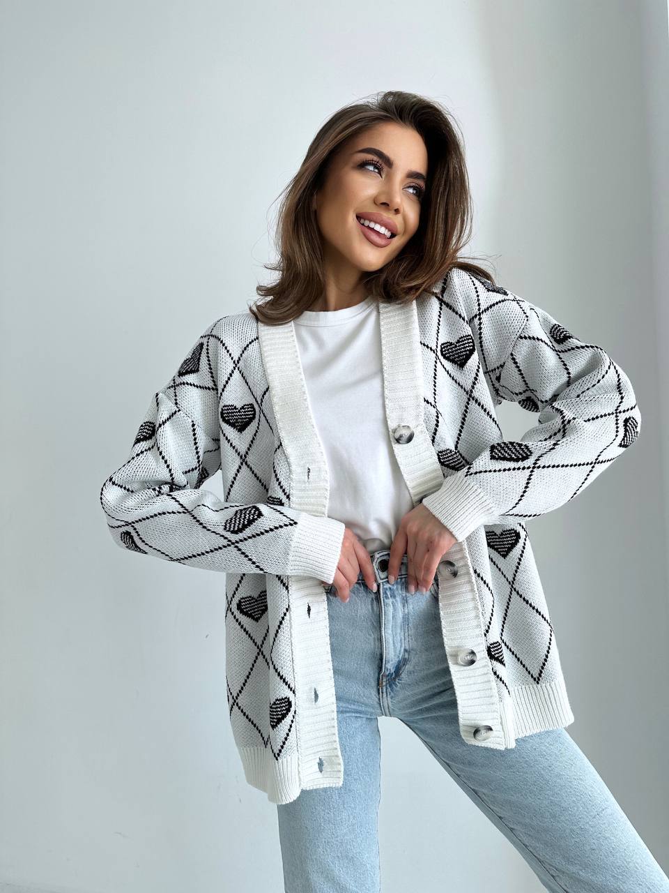Woman wearing a patterned cardigan over a white shirt and light blue jeans on a plain background