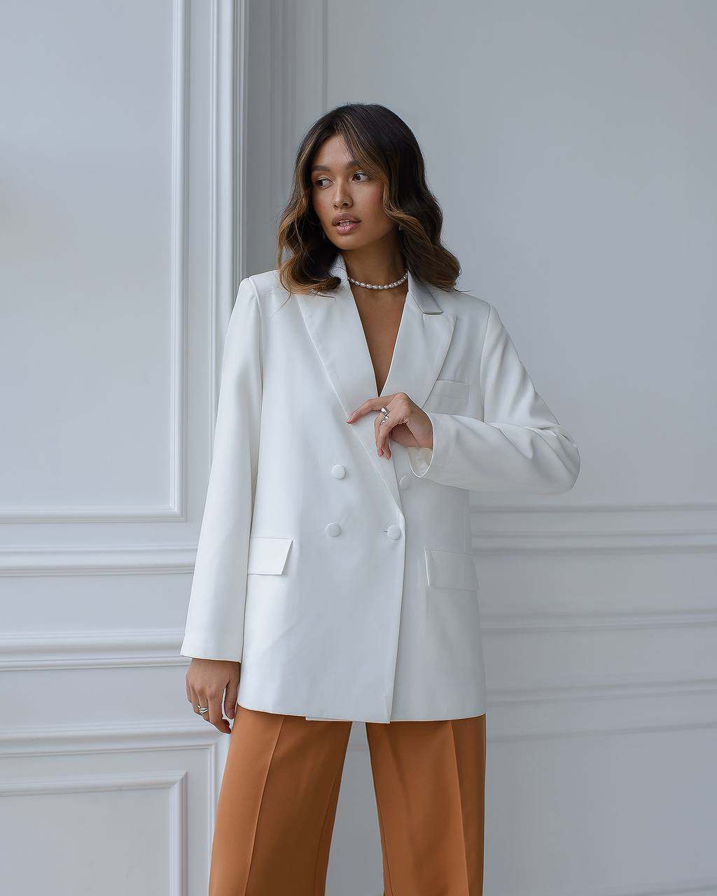 Woman wearing a white blazer and brown pants against a light gray wall.