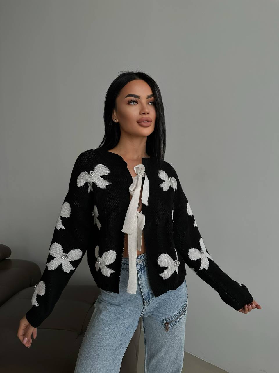 Woman wearing a black cardigan with white floral patterns and light blue jeans against a plain background