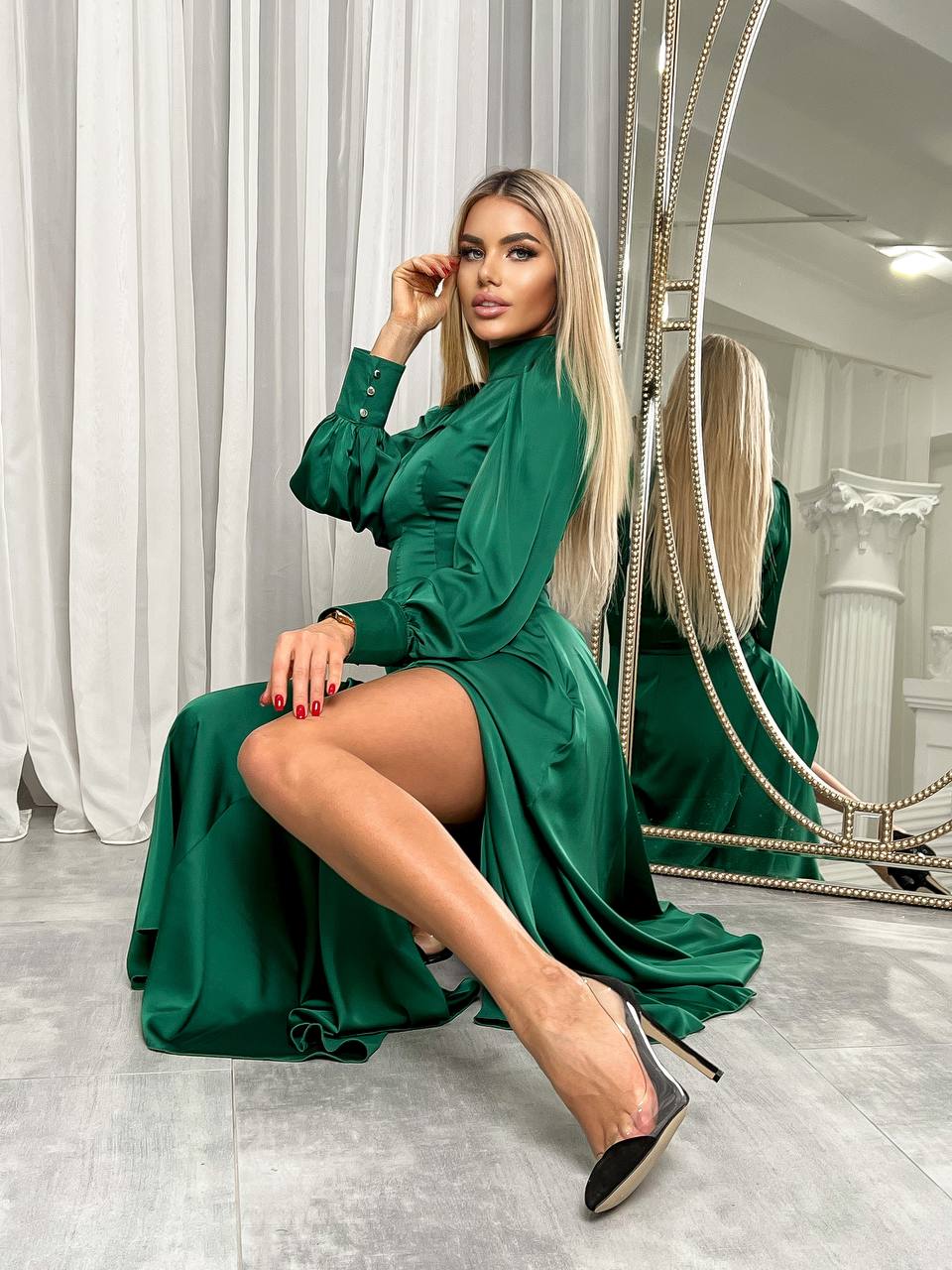 Woman in a green dress sitting in front of a mirror in a room with white curtains.