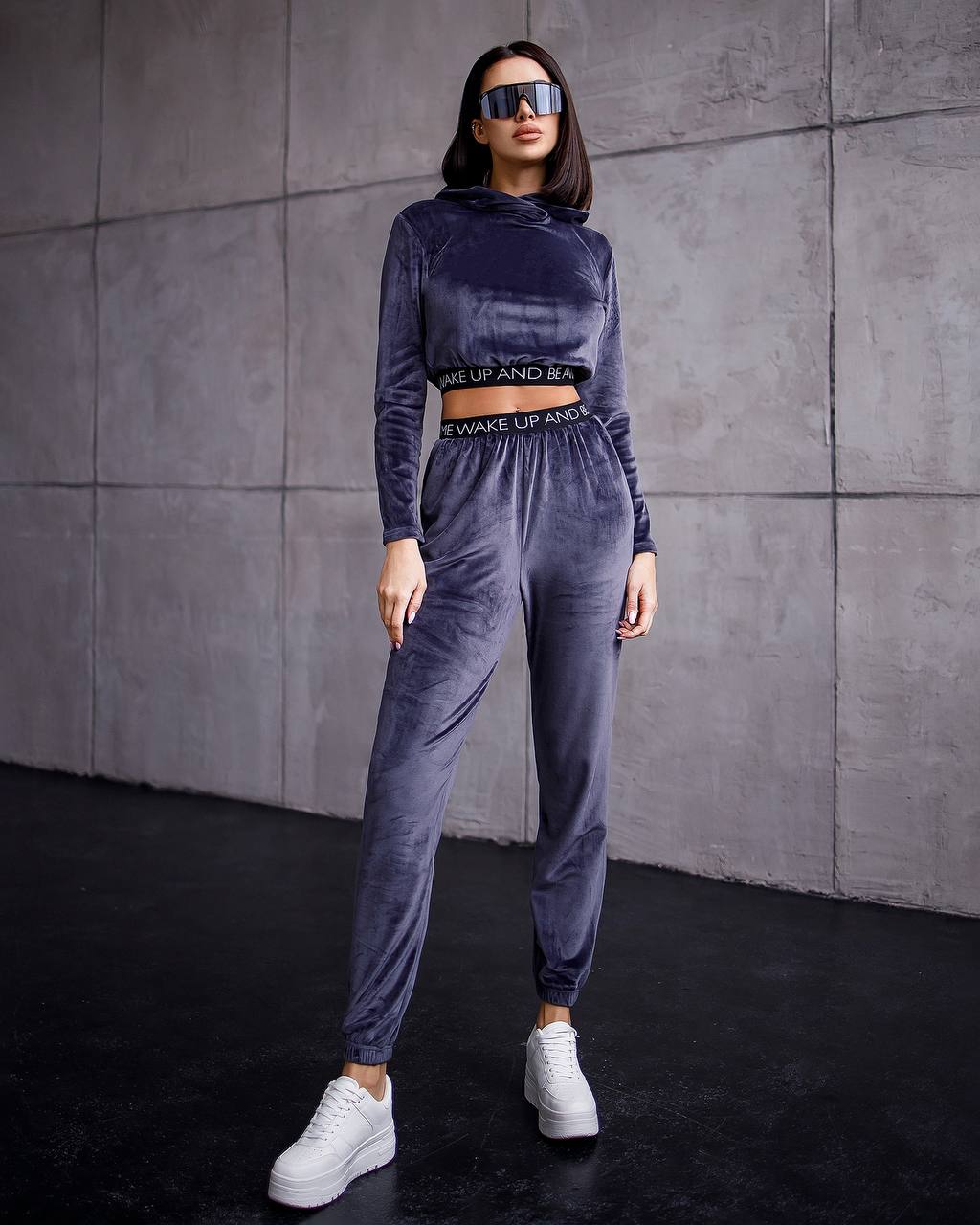 Woman wearing a gray velour tracksuit with text on it against a gray wall.