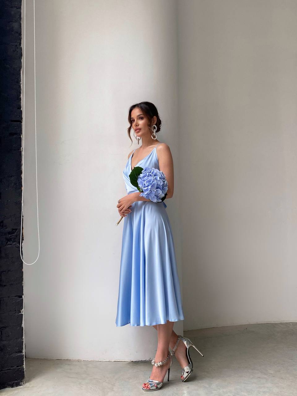 Woman in a light blue dress holding flowers against a plain wall.