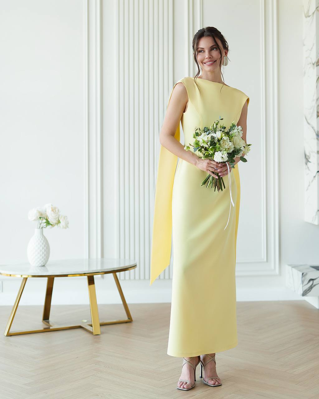 Woman in a yellow dress holding flowers in a room with white walls and a gold table.