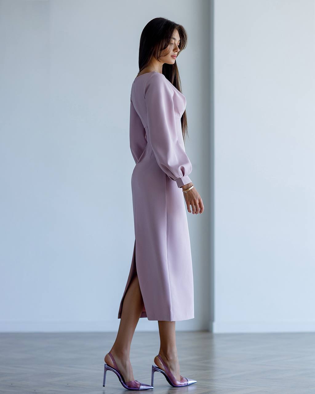 Woman wearing a light purple dress with a high slit, standing against a plain white wall.