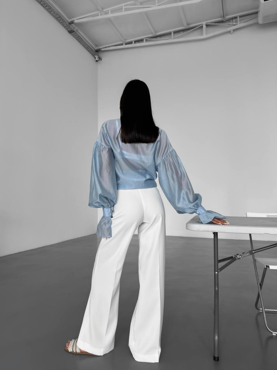 Person wearing a light blue sheer top and white pants in a minimalistic room.