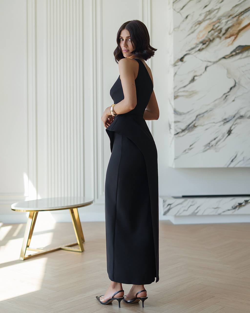 Woman in a black dress standing in a stylish room with marble wall and gold table.