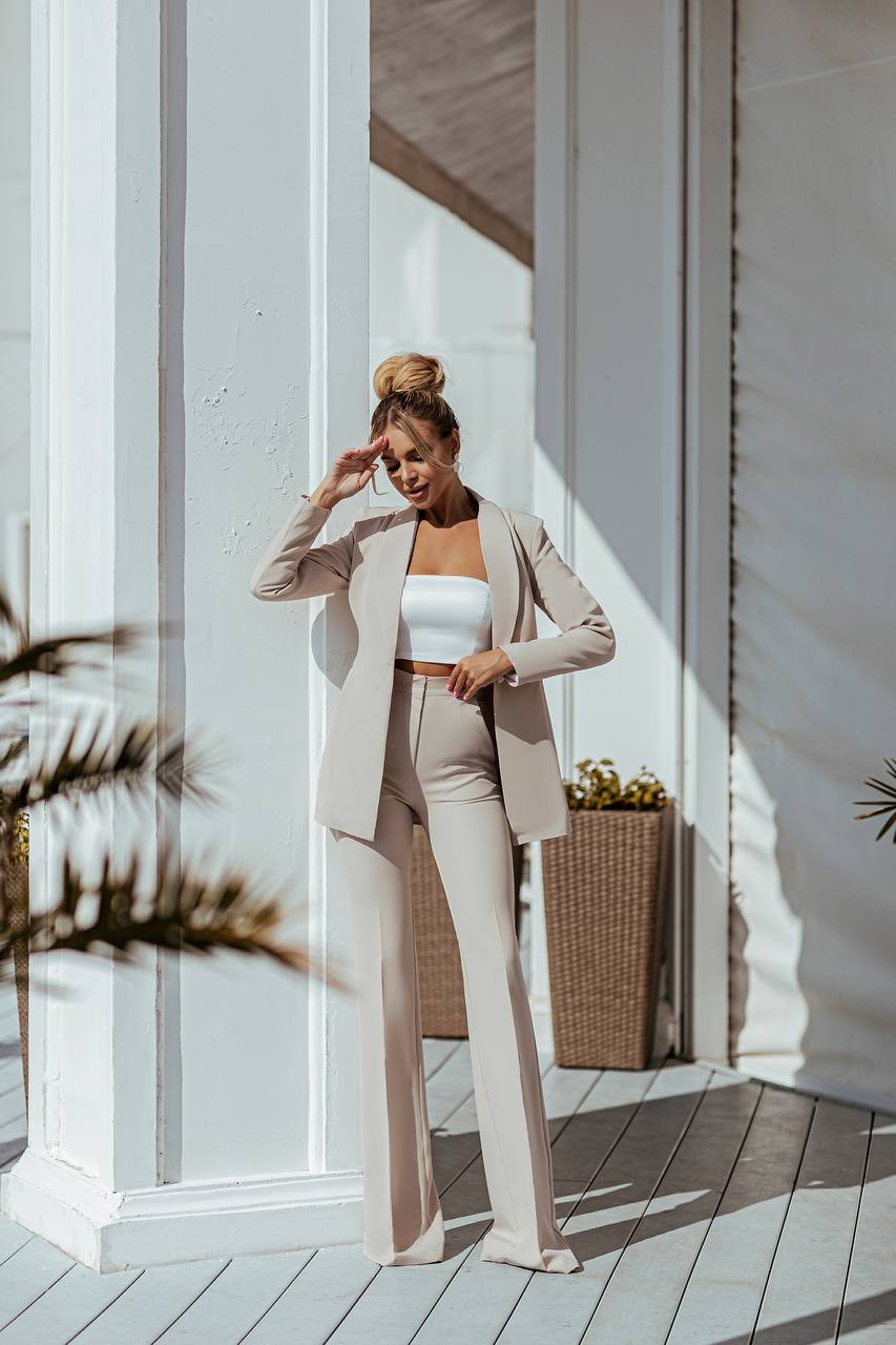 Woman in a beige suit standing in a sunlit room with white walls and wooden floor.