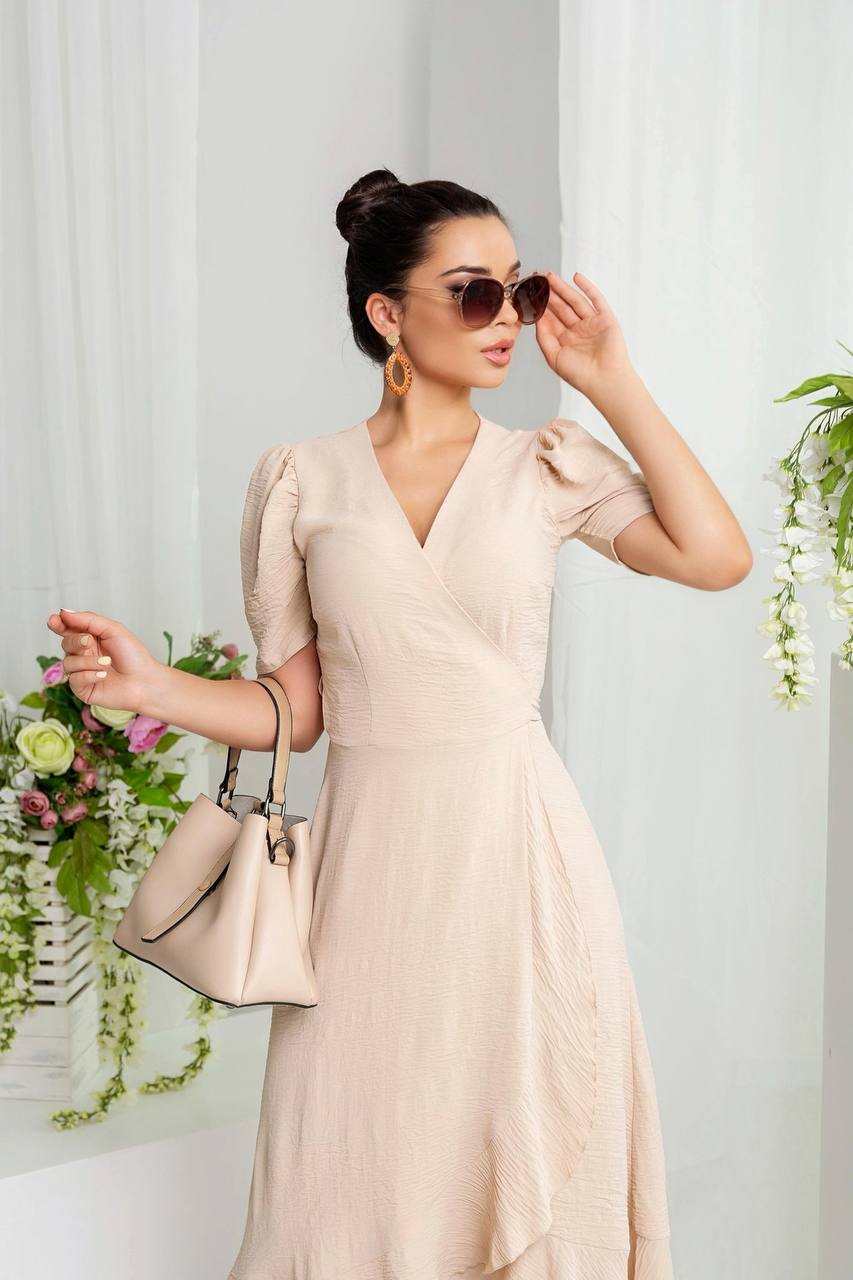 Woman in a beige dress with sunglasses and handbag against a white curtain background