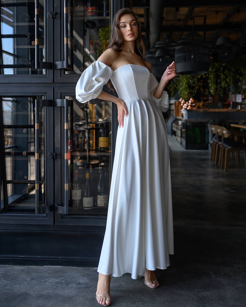 Woman in a white off-shoulder dress standing in a dimly lit room with dark wood cabinets.