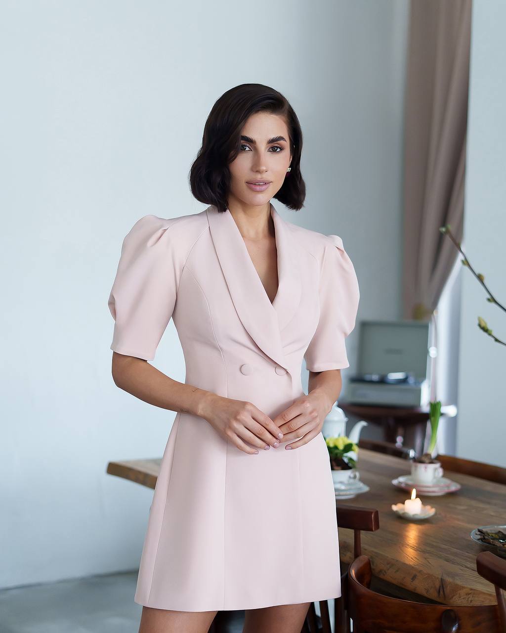 Woman in a light pink dress standing in a room with a table and decor.